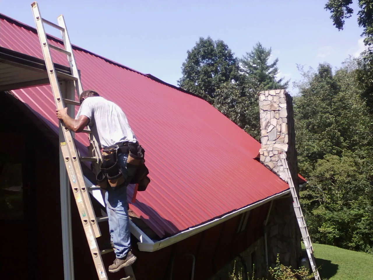 Roofer on a ladder beside a red metal roof and stone chimney — Soffit & Fascia Repair by Roofing Brothers in Omro